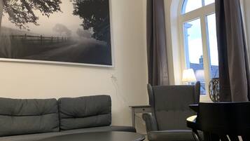 Ein Wohnzimmer mit einer Couch, einem Sessel, einem Tisch, einem Gemälde und einem Fenster mit Vorhängen.