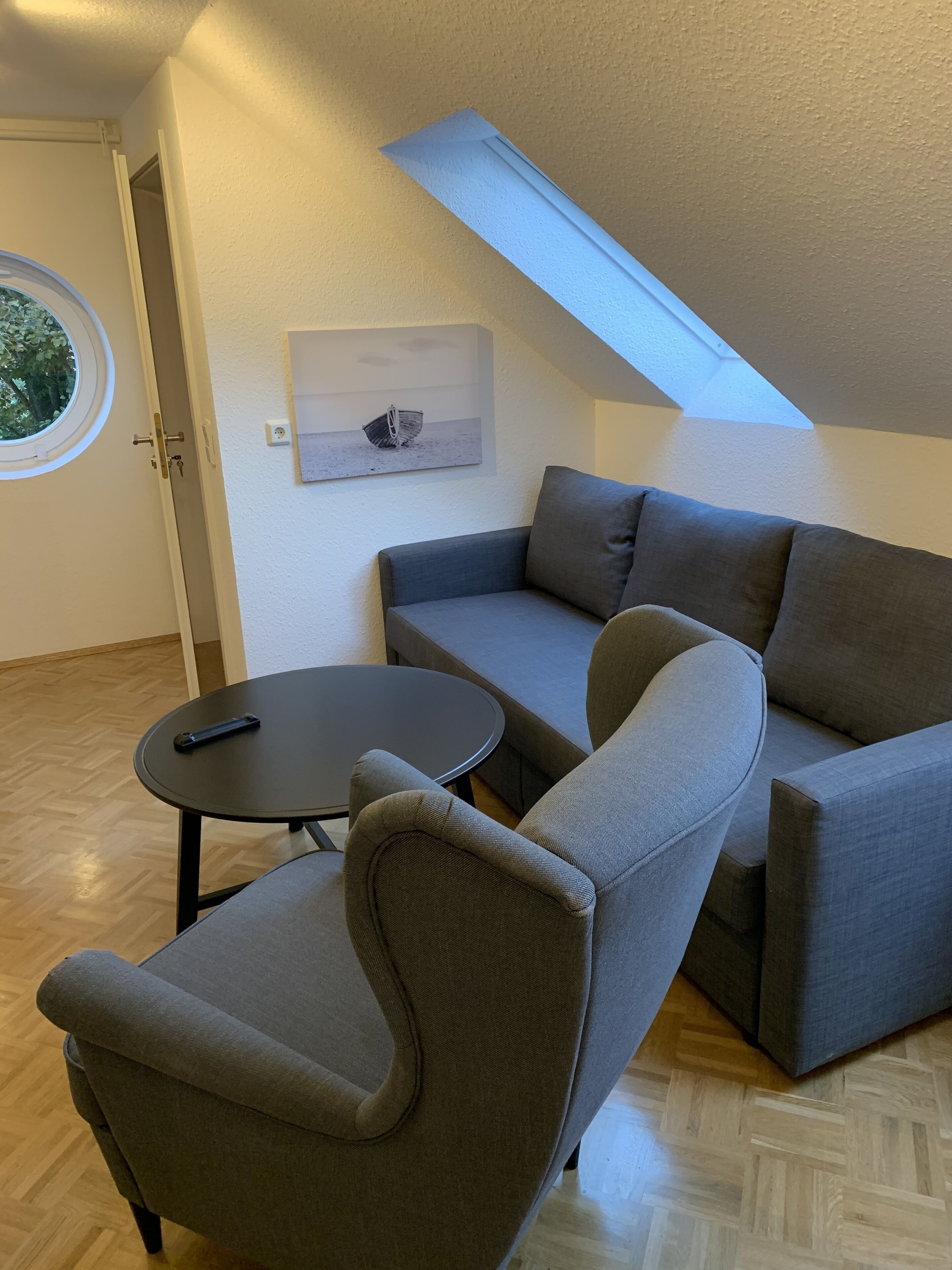 A modern living room with a gray sectional sofa, a round coffee table, and a small armchair. A skylight is visible above the sofa, and a painting hangs on the wall.