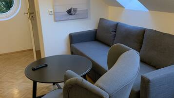 A modern living room with a gray sectional sofa, a round coffee table, and a small armchair. A skylight is visible above the sofa, and a painting hangs on the wall.