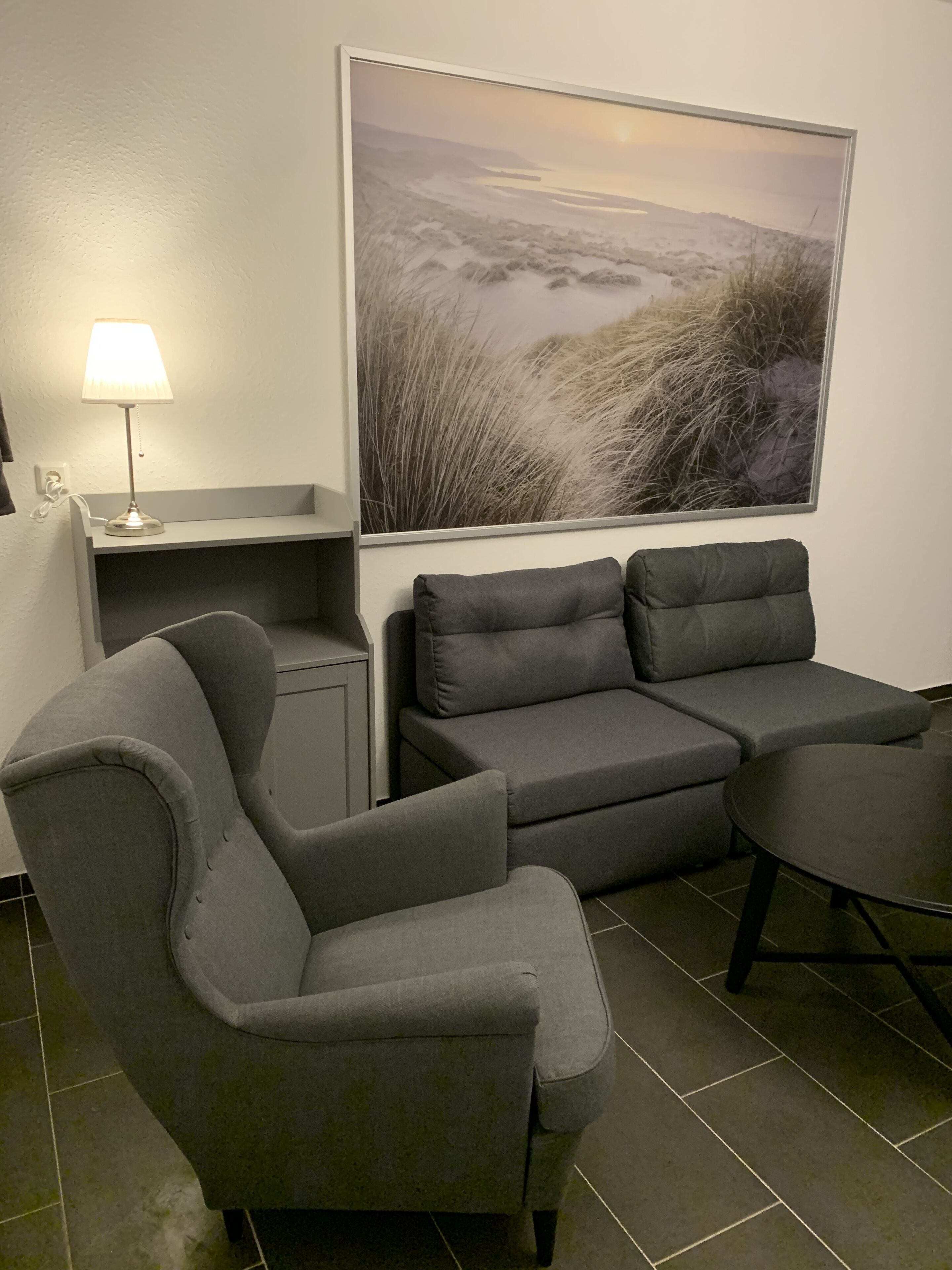 A modern living room with a grey sofa set, a small table, and a lamp. A large landscape painting is on the wall.