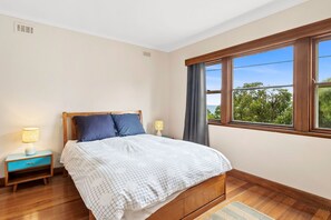 A bedroom with a wooden bed, two bedside tables with lamps, a window with curtains, and a view of greenery outside. - Smith (Lorne)