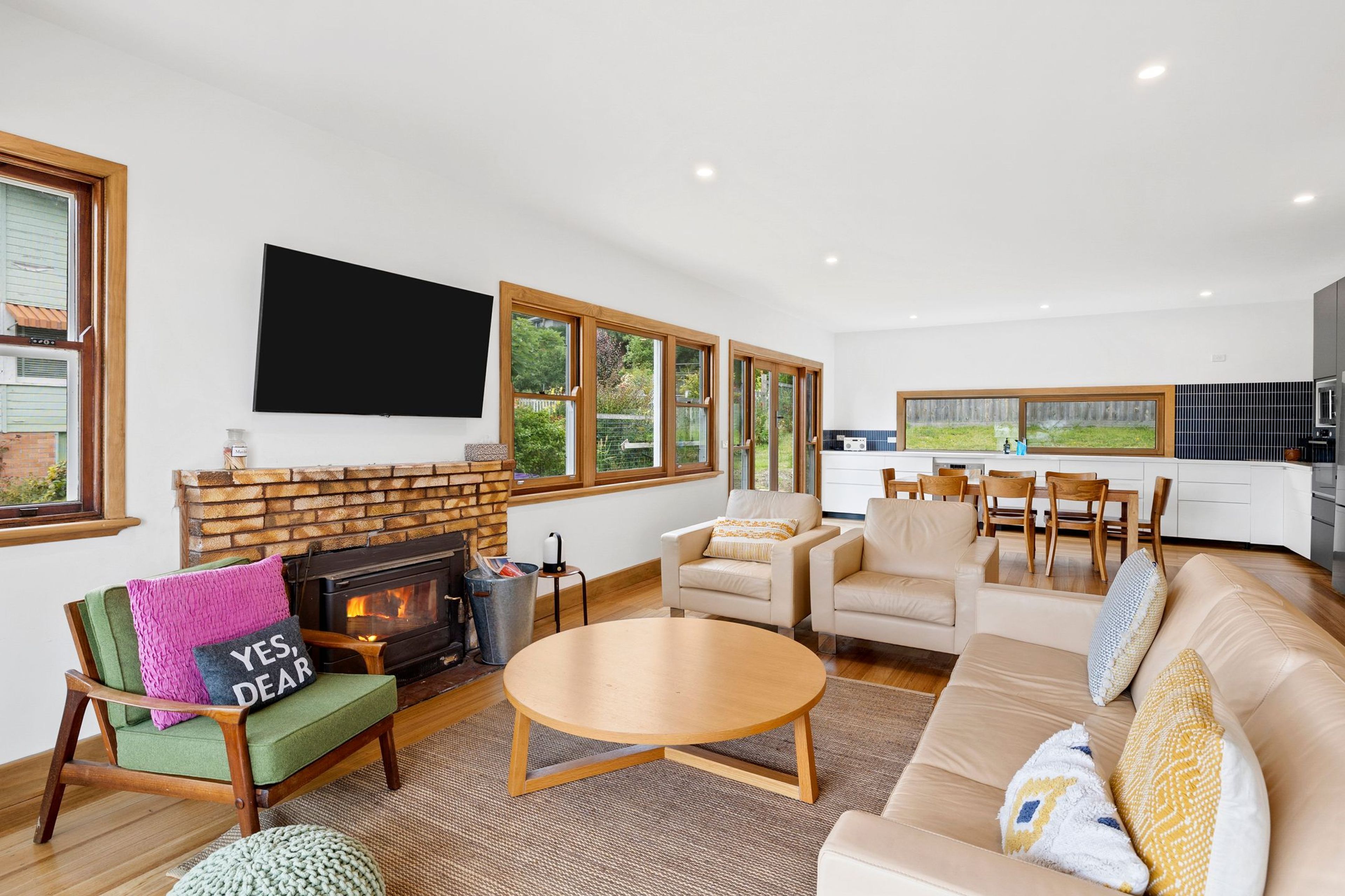 A modern living room with a fireplace, a flat-screen TV, and a dining area with a kitchen in the background.
