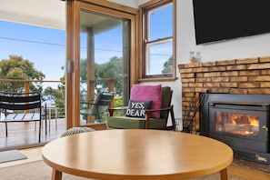 A cozy living room with a round wooden table, a green chair with a cushion, a fireplace, and a balcony with a wooden railing.