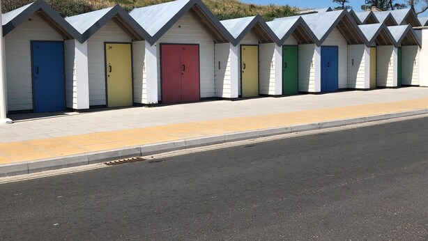 A row of beach huts with numbered doors, a clear sky, and a grassy hill in the background.