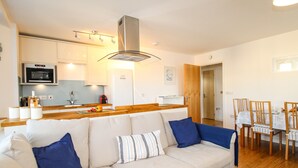 A modern living room with a beige sofa, blue cushions, a wooden coffee table, and a kitchen area with a microwave, oven, and a wooden cabinet.
