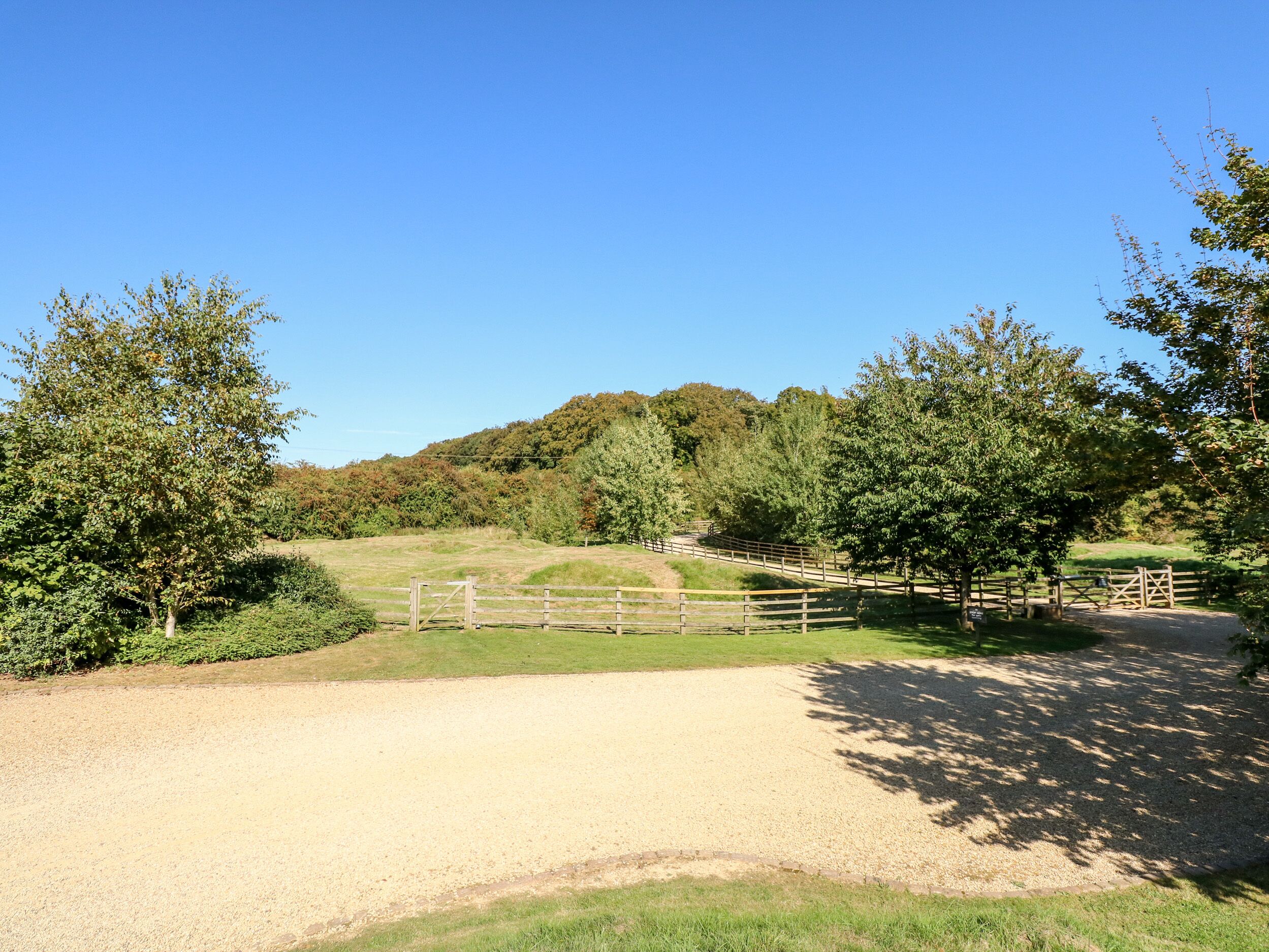A gravel path leading to a wooden gate, surrounded by trees and a grassy field.