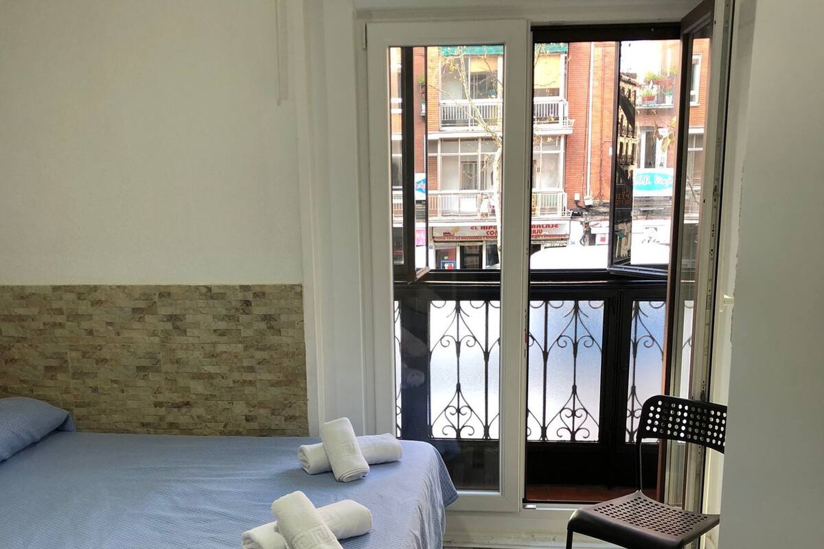Habitación de hotel con cama, una silla y vista a la calle por una puerta abierta.