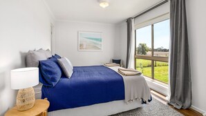 A bedroom with a large window, a bed with blue bedding, a bedside table with a lamp, and a painting on the wall. - Bass River Farm 9 mins from Phillip Island (Bass)
