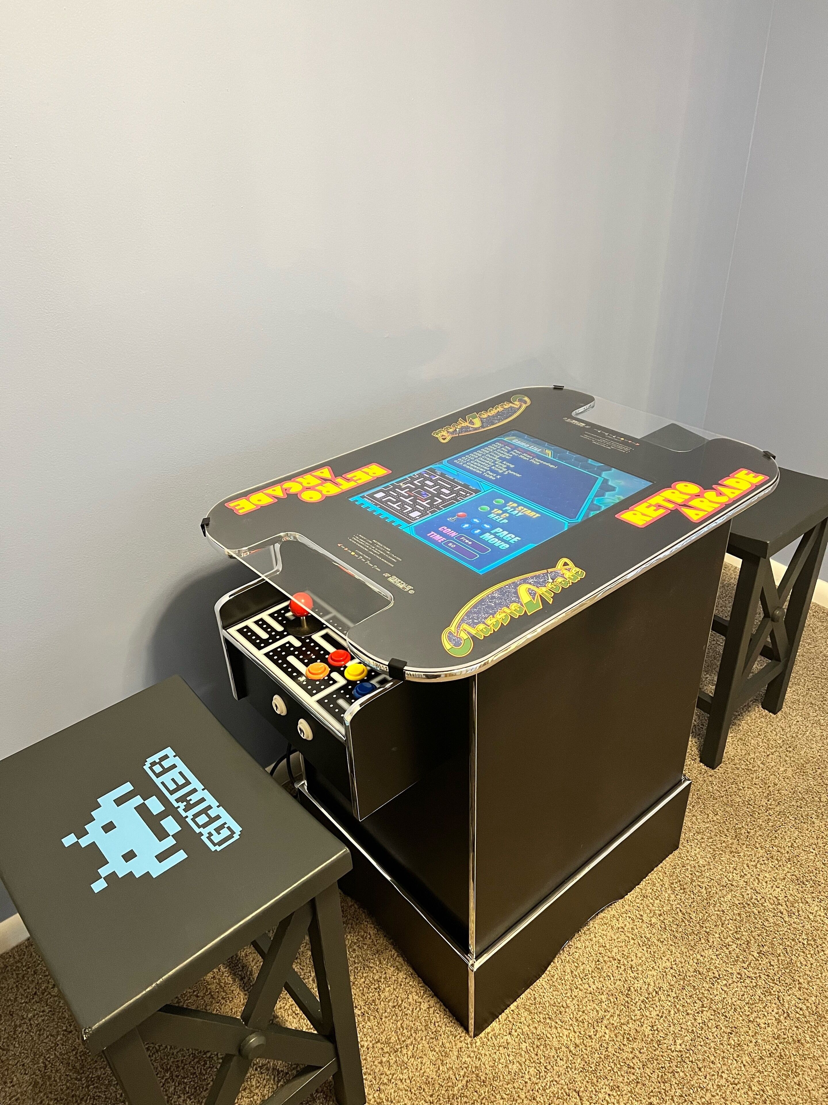 A retro arcade table with a joystick and buttons, a small table with a game controller, and a wall-mounted screen displaying game options.
