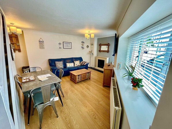 A living room with a blue sofa, a wooden coffee table, a dining table with chairs, a fireplace, and a window with blinds. - A perfect location for a Tenby break (Tenby)