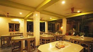 A restaurant with multiple tables and chairs, ceiling fans, and potted plants. - Neel Nirjan Resort (Bolpur)