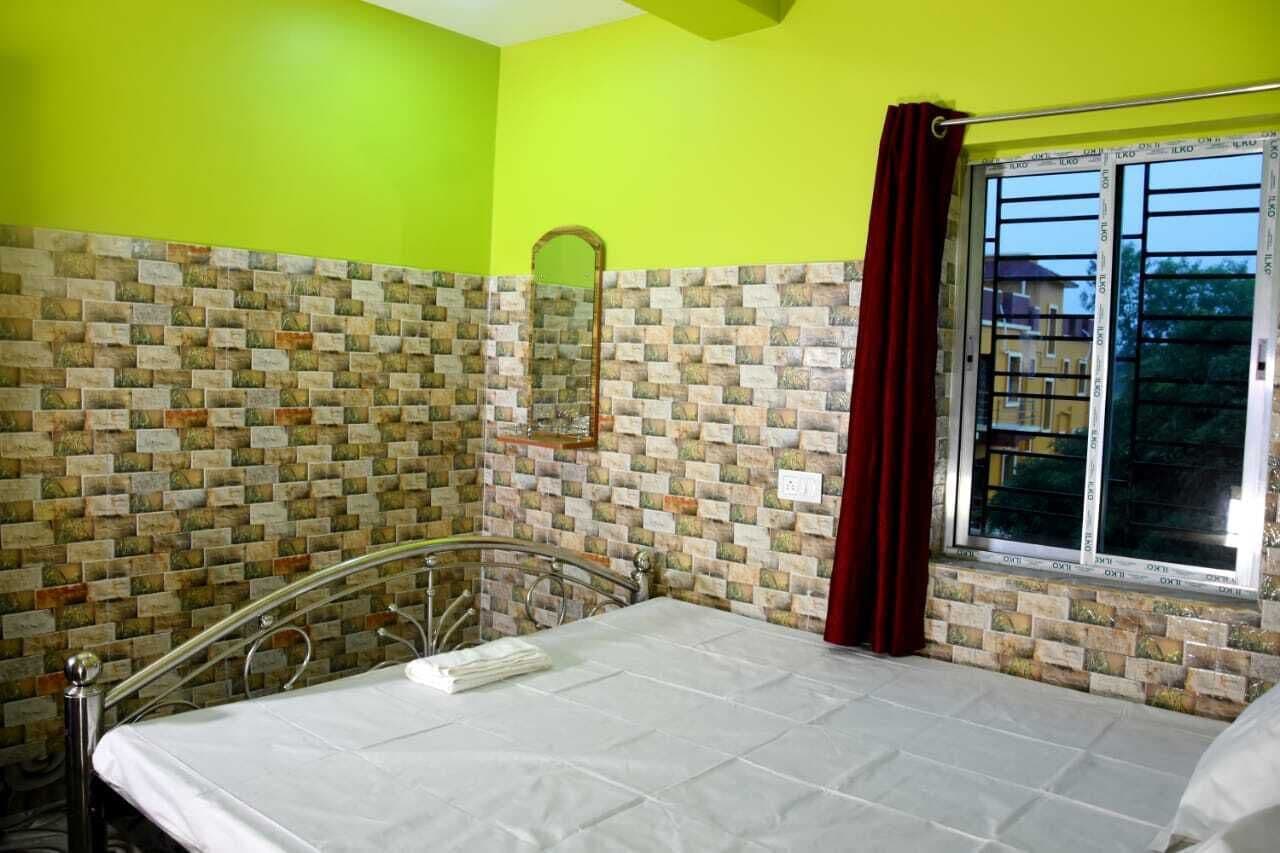 Una stanza con un letto, una sedia con struttura in metallo, una finestra con tenda e un muro in mattoni.