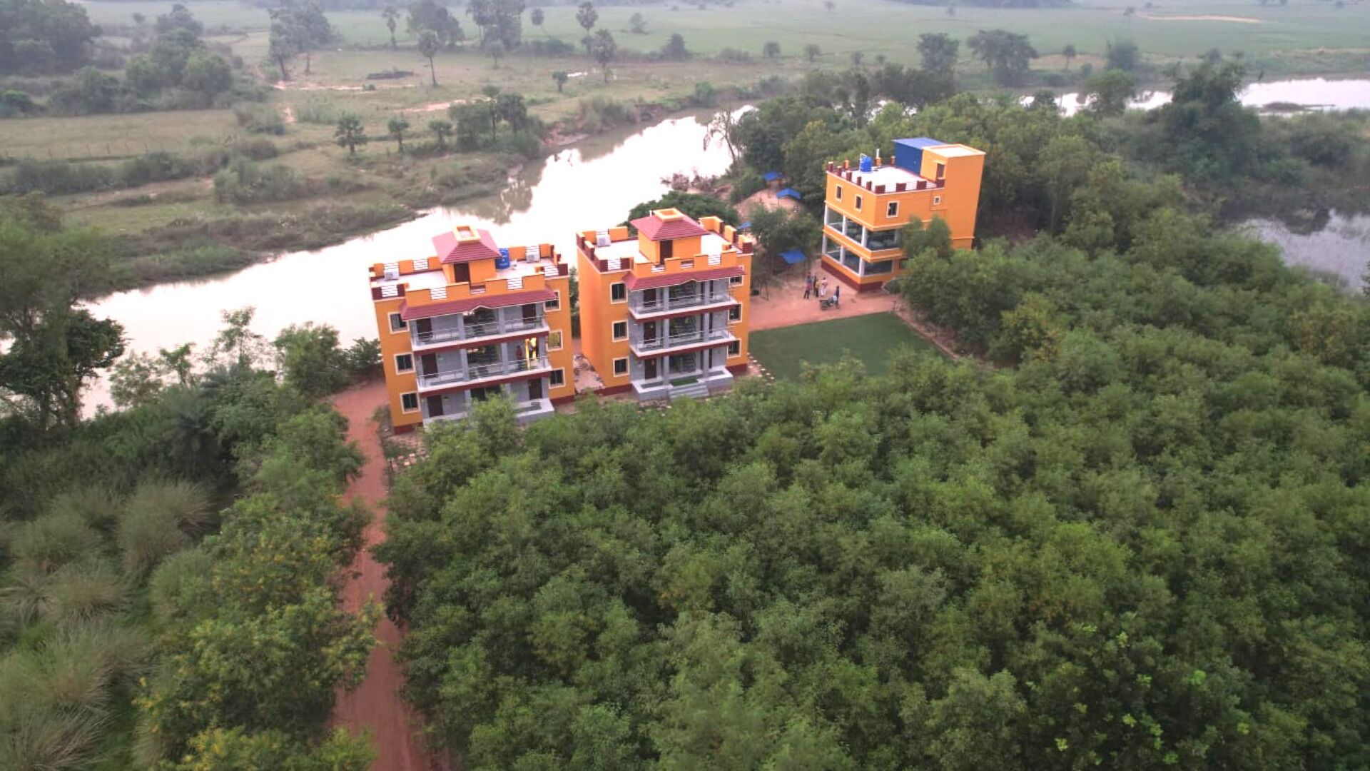 A multi-story hotel with a flat roof, surrounded by dense greenery and a river.