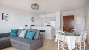 Een moderne woonkamer met een grijze hoekbank, een eettafel met witte stoelen, en een keuken met witte kasten en een houten deur.