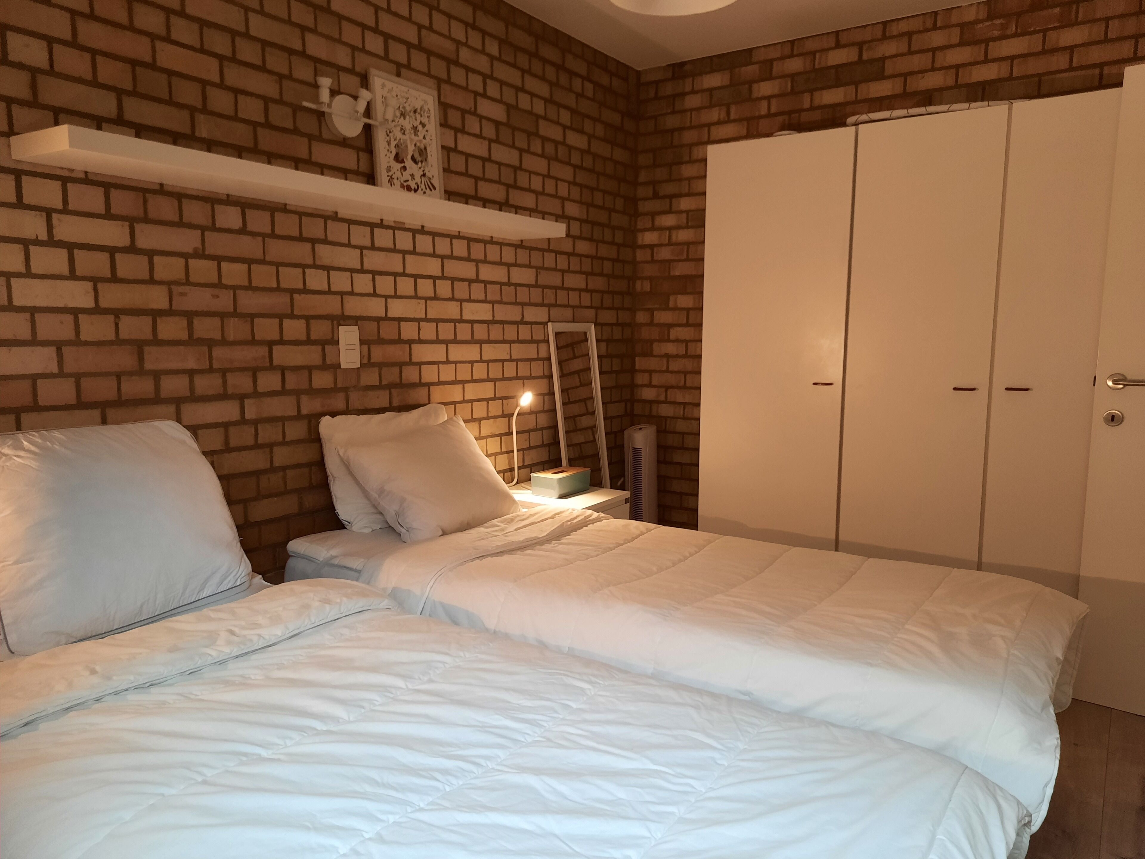 A bedroom with a brick wall, a white bed, a bedside table with a lamp, and a wardrobe.