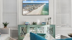 A living room with a large TV mounted on the wall displaying a beach scene, a teal cabinet with decorative items, and a cozy seating area with cushions. - Steps to beach, BBQ, bikes, pool, arcade, Wifi (Gulf Shores)