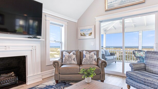 A living room with a fireplace, a large flat-screen TV, a sofa with patterned cushions, and a view of the outdoors through large sliding glass doors. - Our most Popular Rental on Tybee! Voted best view! by Tybee Cottages (Tybee Island)