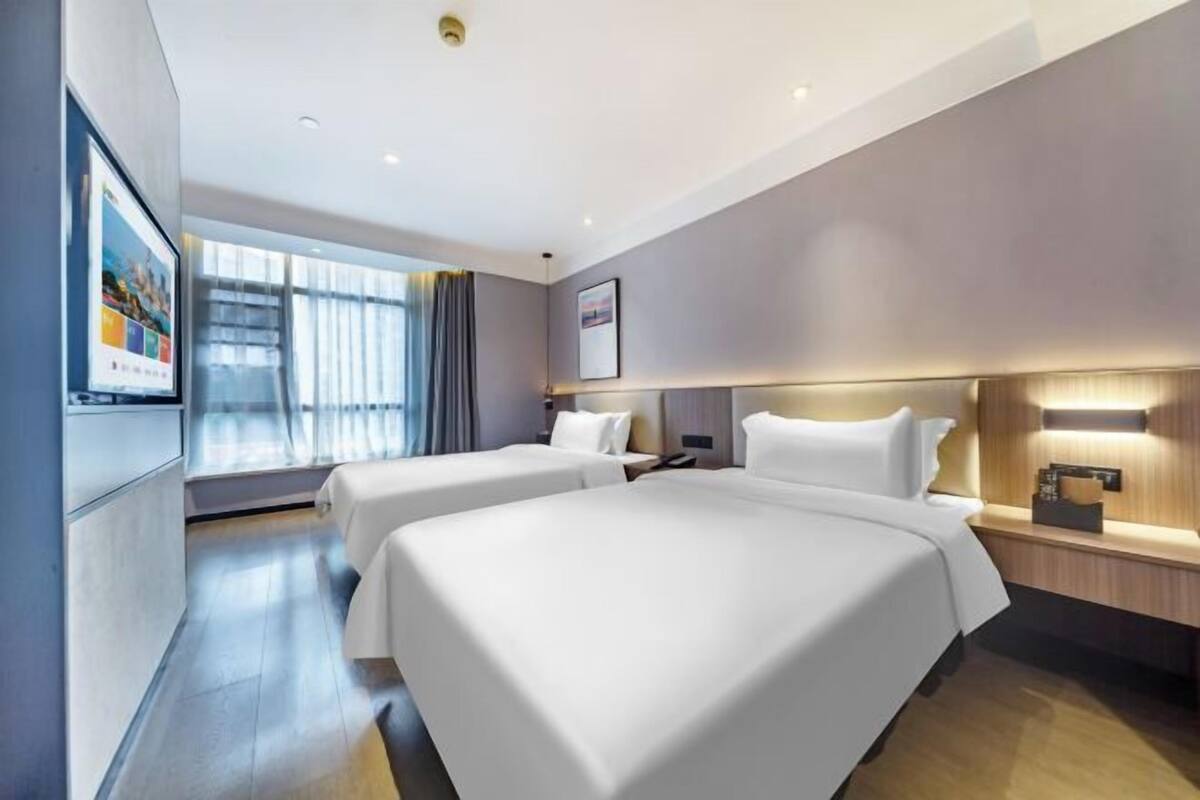 Une chambre d’hôtel moderne avec deux lits, une grande fenêtre avec des rideaux, un téléviseur à écran plat et une table de chevet en bois.