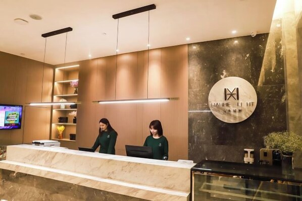 A hotel reception area with a marble counter, two receptionists, a television, and a wall-mounted logo. - Wahme Light Hotel (Shangrao)