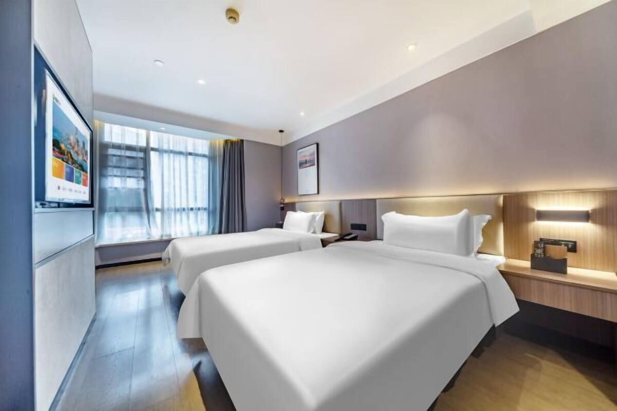 Une chambre d’hôtel moderne avec deux lits, une grande fenêtre avec des rideaux, un téléviseur à écran plat et une table de chevet en bois.