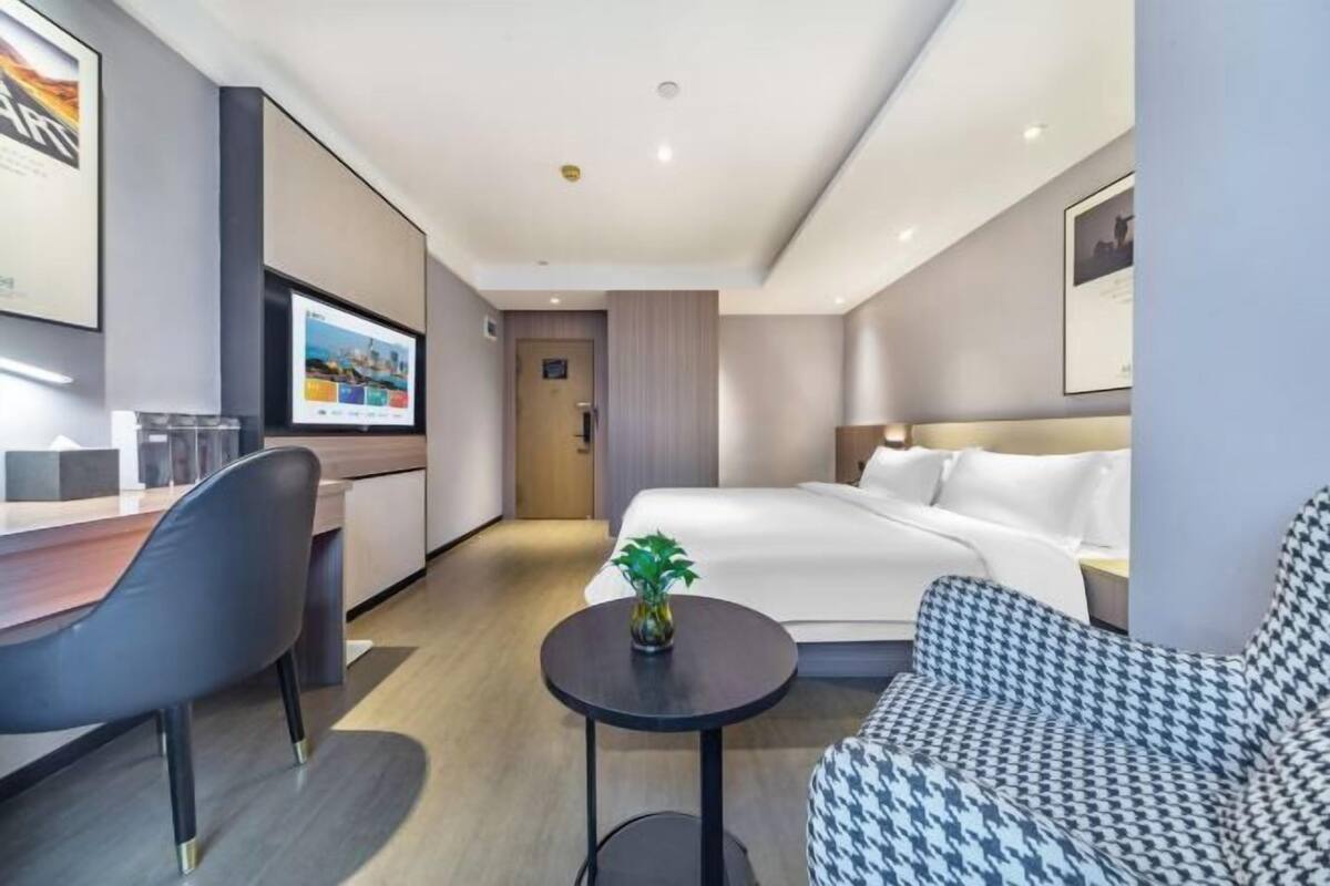 Une chambre d’hôtel moderne comprenant un lit, un bureau, une chaise, une petite table avec une plante et un canapé à motifs pied-de-poule.