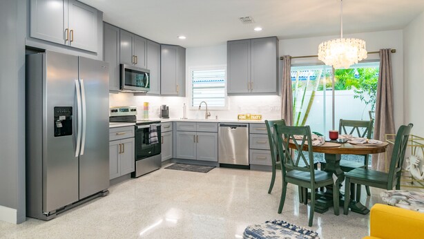 A modern kitchen with grey cabinets, stainless steel appliances, and a dining area with a yellow sofa and green chairs. - Made in Shade B · Beach Across St · Pets OK (Holmes Beach)