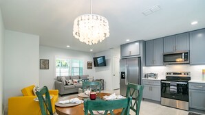 A modern kitchen with grey cabinets, a stainless steel refrigerator, and a microwave. There is a dining area with a wooden table and green chairs. - Made in Shade B · Beach Across St · Pets OK (Holmes Beach)