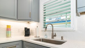 A modern kitchen with grey cabinets, a stainless steel sink, and a window with blinds. - Made in Shade B · Beach Across St · Pets OK (Holmes Beach)