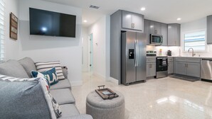A modern kitchen with grey cabinets, a stainless steel refrigerator, a microwave, and a dishwasher. There is a grey sofa with striped pillows in the living area. - Made In The Shade A &B | Multiple Units (Holmes Beach)