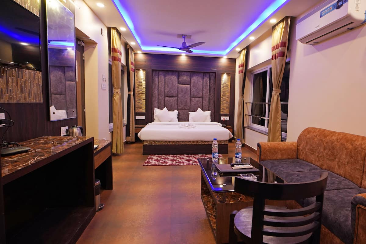 A hotel room with a bed, a sofa, a desk with a chair, a mirror, and an air conditioning unit.