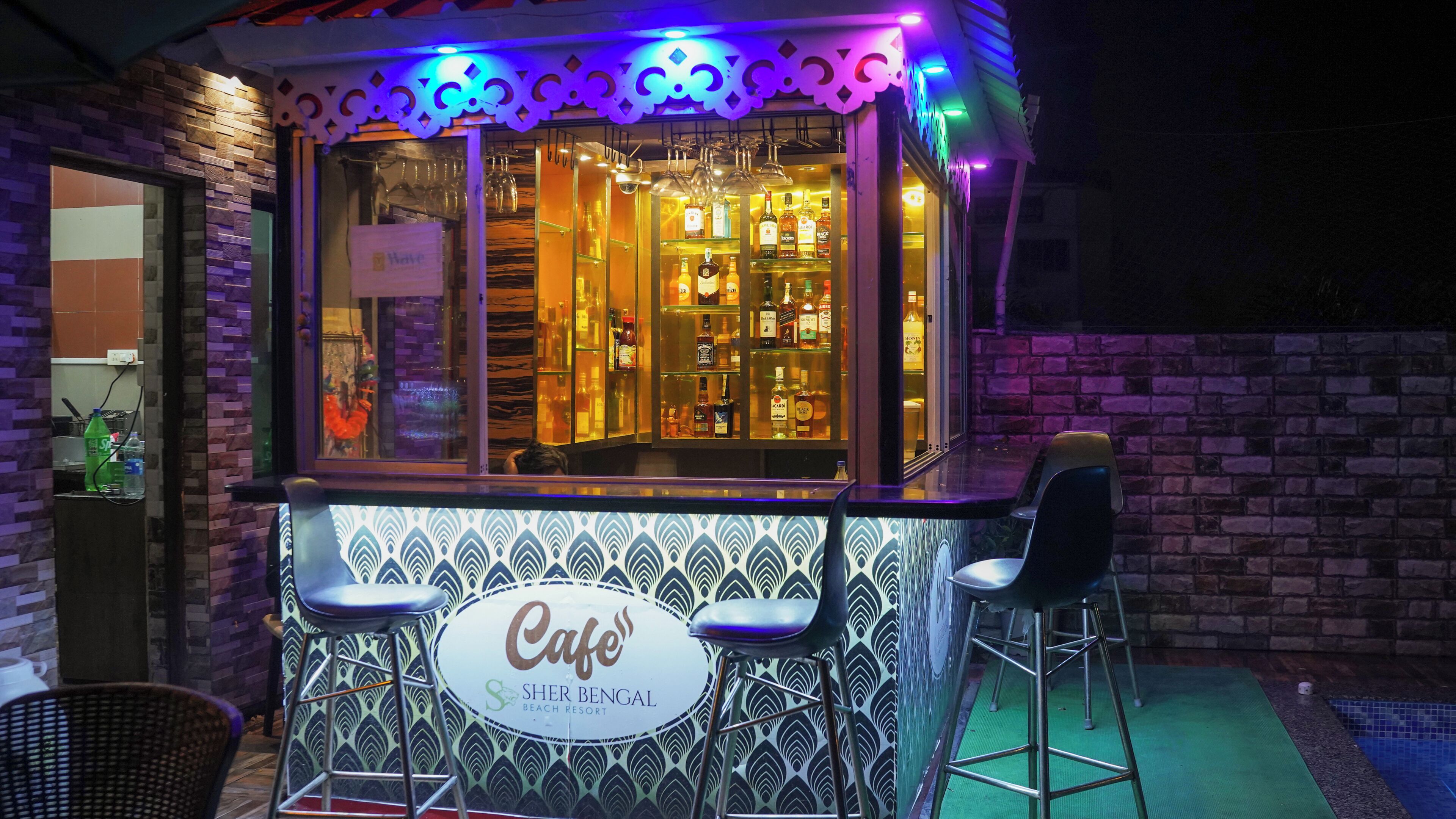 A bar with a patterned exterior, stools, and a sign reading 'Café Sher Bengal Beach Resort.'