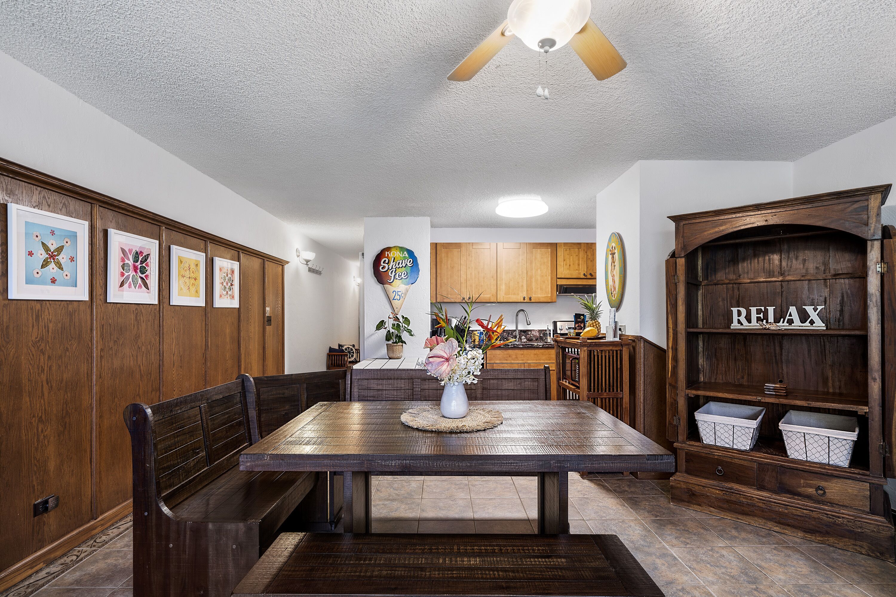 Un espace repas avec une table et des bancs en bois, un ventilateur de plafond et une cuisine en arrière-plan.