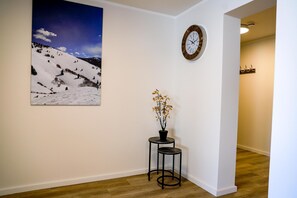 A hallway with a wooden floor, a wall-mounted clock, a framed picture of a snowy landscape, and a small table with a potted plant.
