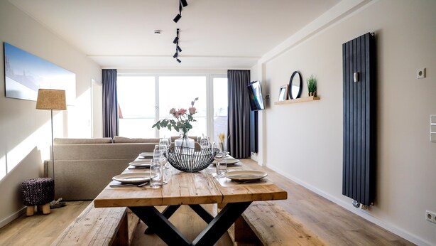 Un coin repas avec une table en bois dressée pour quatre personnes, des bancs de chaque côté, un vase avec des fleurs et un canapé en arrière-plan.