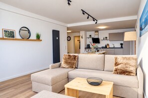 Una sala de estar moderna con un sofá modular, una mesa de centro de madera y una cocina al fondo.