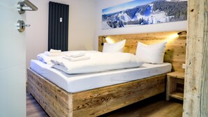 Un lit en bois avec du linge de lit blanc, un radiateur noir et, au-dessus, un tableau représentant un paysage de montagne.