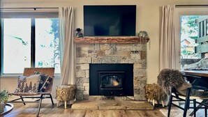 Un salón con chimenea de piedra, un televisor de pantalla plana montado encima y un amplio ventanal con cortinas.