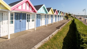 Une rangée de cabanes de plage colorées, dotées de portes et de fenêtres blanches, un chemin de gravier et une haie bien entretenue le long de la plage.