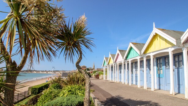 Une rangée de cabanes de plage colorées, dotées de piliers blancs et de portes bleues, un palmier et un ciel bleu azur.