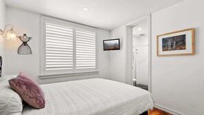 A bedroom with a bed, white walls, a window with blinds, a framed picture, and a door leading to a bathroom.