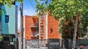A multi-story brick building with a gated entrance, a tree in front, and a parked car.