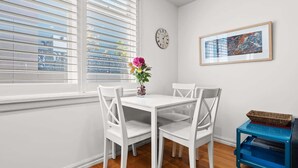 A dining area with a white table and chairs, a vase with flowers, a clock, and a framed artwork on the wall.