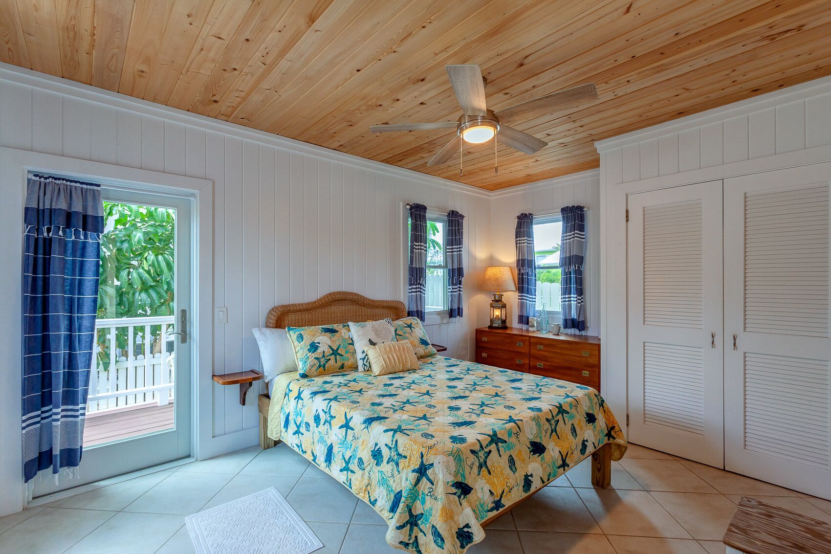 A bedroom with a wooden ceiling, a bed with a floral bedspread, a nightstand, a dresser, and a window with curtains.