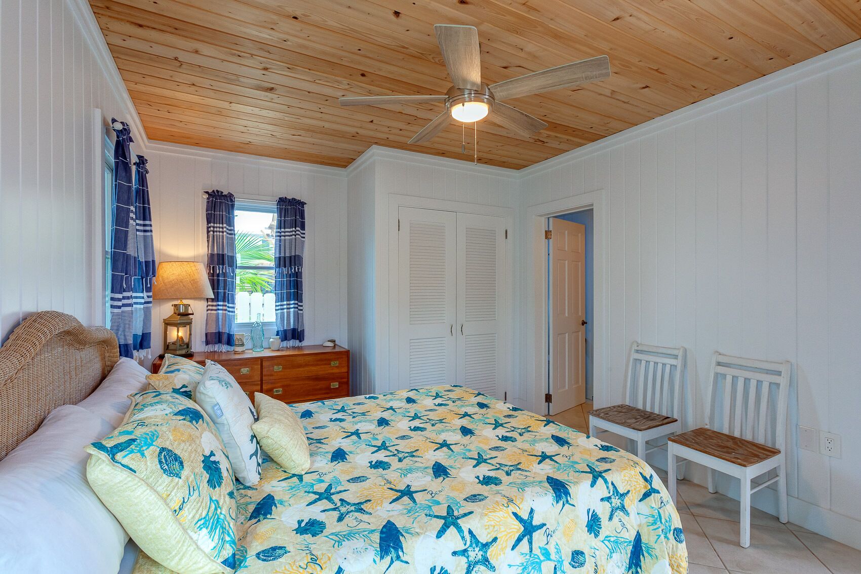A bedroom with a wooden ceiling, a bed with a starfish-themed bedspread, a nightstand with a lamp, a dresser, and two white chairs.