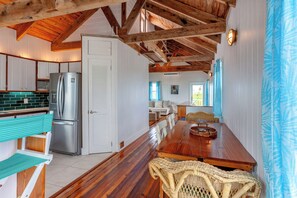 A spacious kitchen with a wooden ceiling, a stainless steel refrigerator, a long wooden dining table, and a wicker chair.