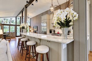 A modern kitchen with a central island, bar stools, and a large window providing a view of the outdoors.
