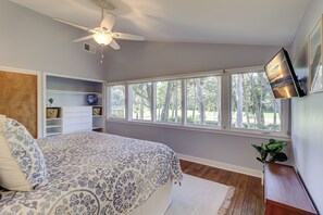 Un dormitorio con cama, ventilador de techo, ventana con vista a árboles, televisor montado en la pared y cabecera de madera.