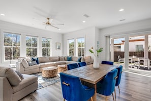 A spacious living room with a large sectional sofa, a wooden dining table with blue chairs, and a ceiling fan.