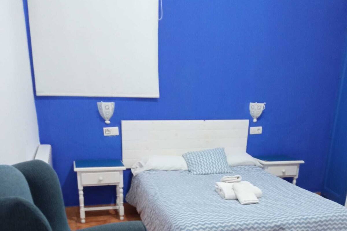 Ein Schlafzimmer mit blauer Wand, einem Bett mit gemustertem Überwurf, einem grünen Sessel und einem weißen Nachttisch mit Lampe.