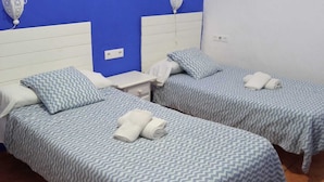 Quarto de hotel com duas camas de solteiro, cada uma com colcha estampada e almofadas, uma mesinha de cabeceira branca e parede azul.